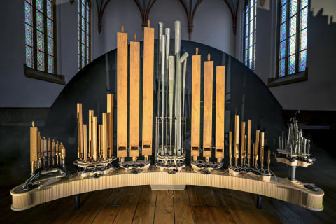 The full instrument on its curved base in a church, wooden and metal pipes rising in front of a dark arched backdrop and stained-glass windows.