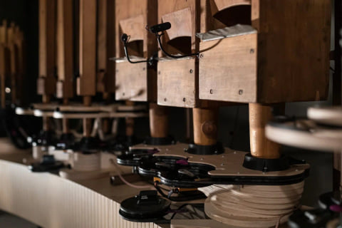 Wooden pipes with small microphones clipped beside their mouths, above robotic mounts on a curved white base.