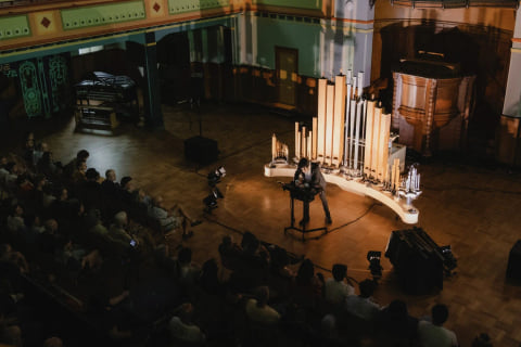 View from a balcony of Navid Navab performing with Organism in a painted hall, the audience seated around the instrument.