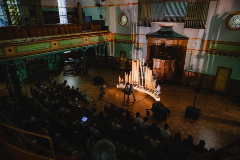A wider view from the balcony of the same hall, with Organism and the performer lit at the centre of the audience.