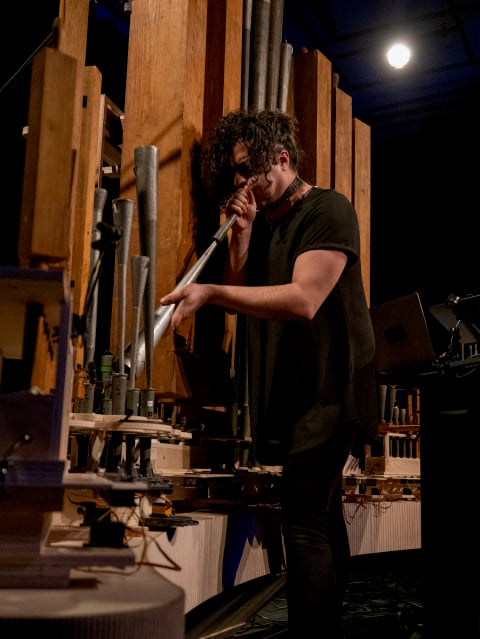 Navid Navab blowing into a metal pipe beside the instrument's wooden pipes.