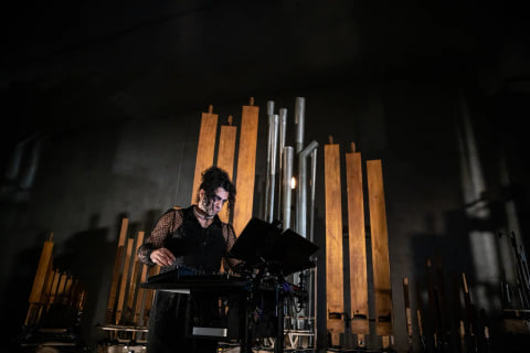 A performer at a control desk in front of the instrument's wooden and metal pipes on a dark stage.