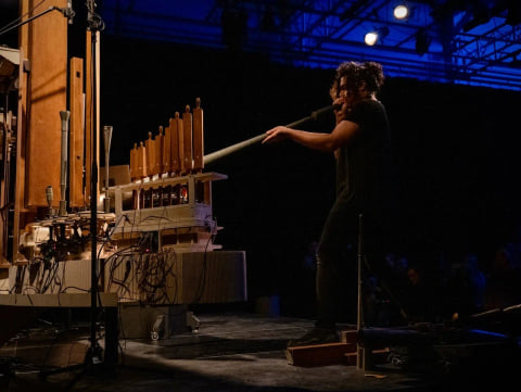 Navid Navab blowing into a long pipe beside the instrument, on a stage lit in blue.