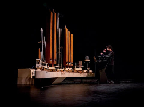 Organism on a long white base on a dark stage, with a performer at a control desk to one side.