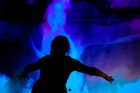 A silhouetted figure with outstretched arms in front of a blue fluid projection in IL Y A.