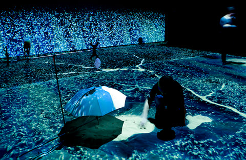 Visitors in a darkened room whose floor is covered in a shimmering blue particle projection, in Einstein's Dream.